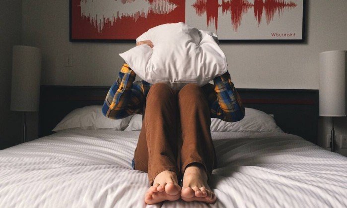 Foto de uma pessoa sentada em cima de uma cama com um travesseiro na cabeça para a pauta "Briga entre vizinhos pode acabar em tragédia" para o Blog da Estasa.