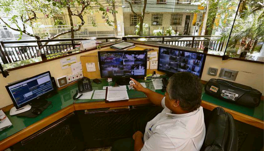 Foto de um porteiro em frente a monitores dentro da portaria de um condomínio para a pauta Ideias para reduzir custos em condomínios para o Blog da Estasa